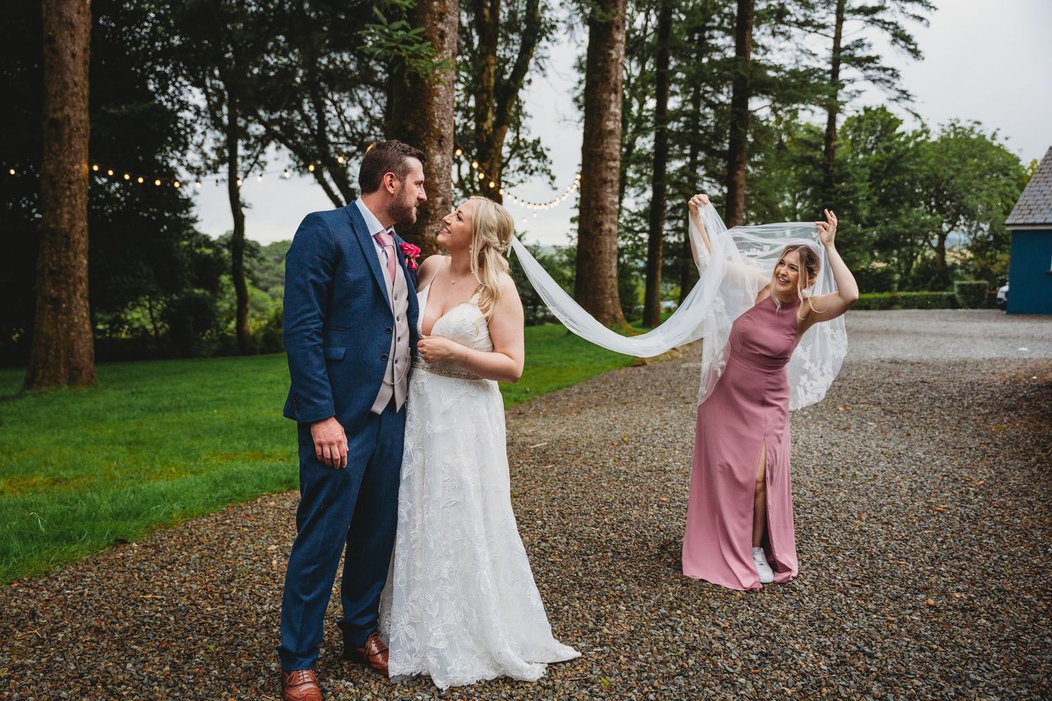 Wedding party celebrating on gravel path with bride's veil flowing in the breeze.