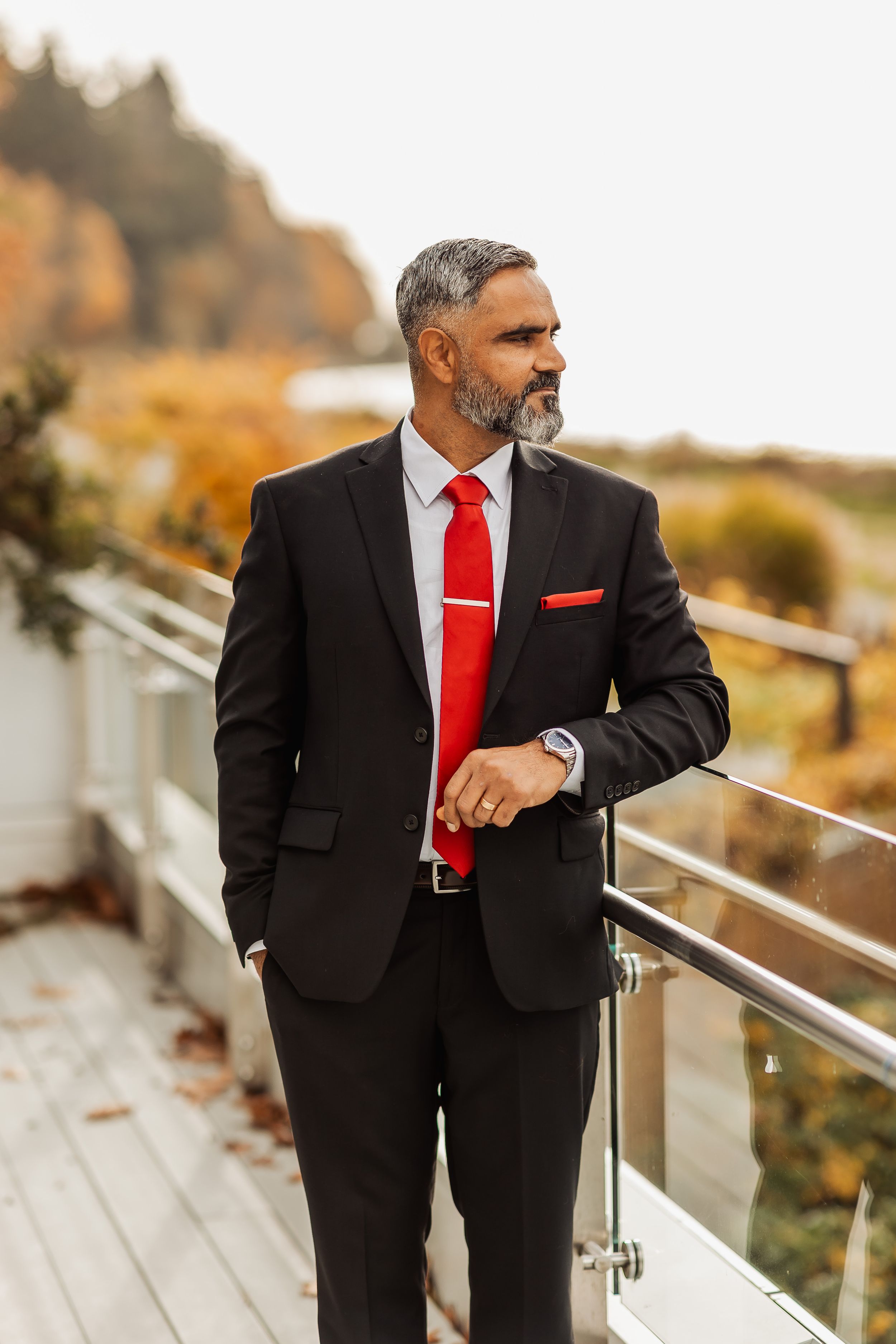 Groom wearing a black suit with red tie leans on railing during autumn outdoor photo session.