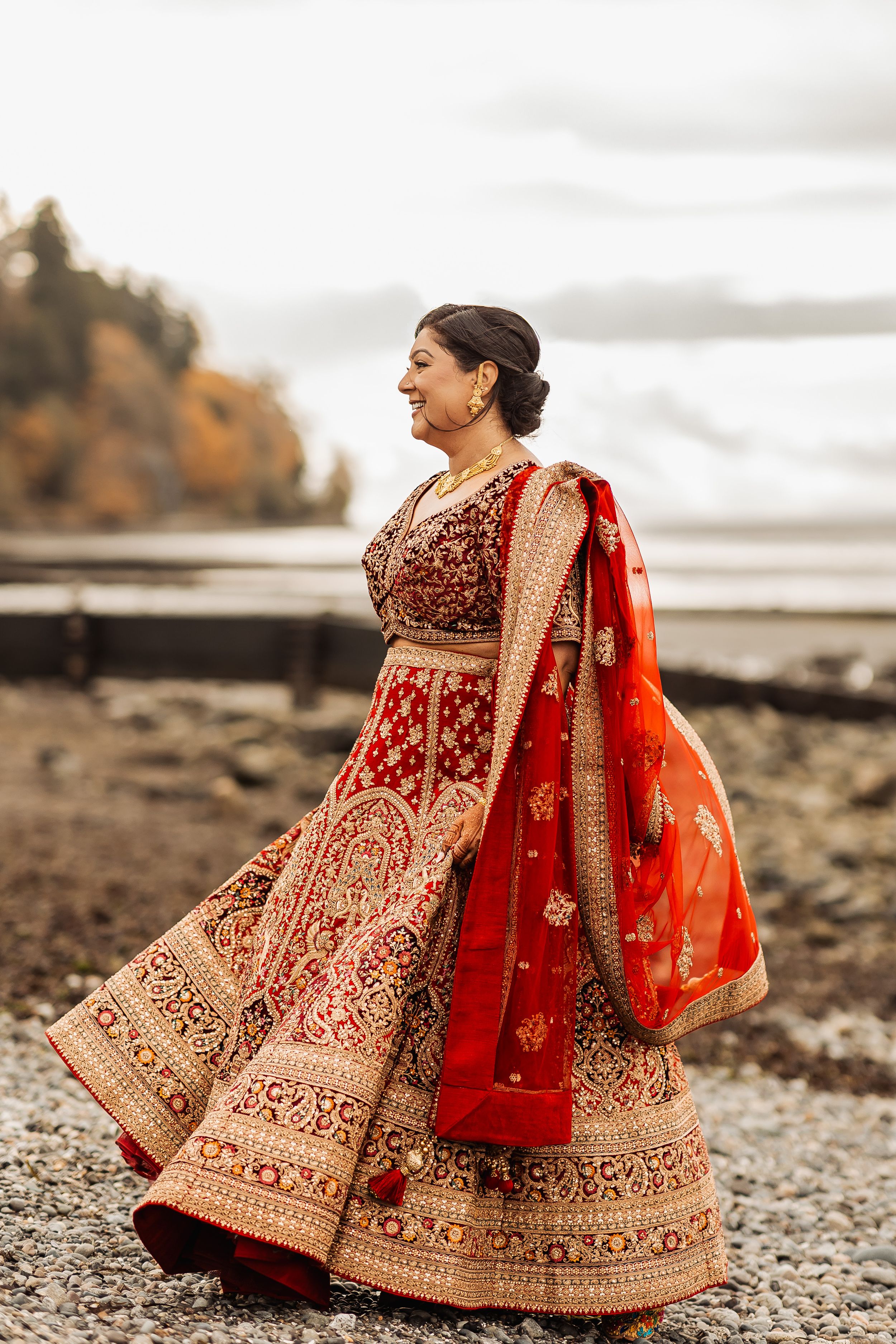 A bride in a traditional red and gold embroidered lehenga poses gracefully on a rocky beach at sunset.