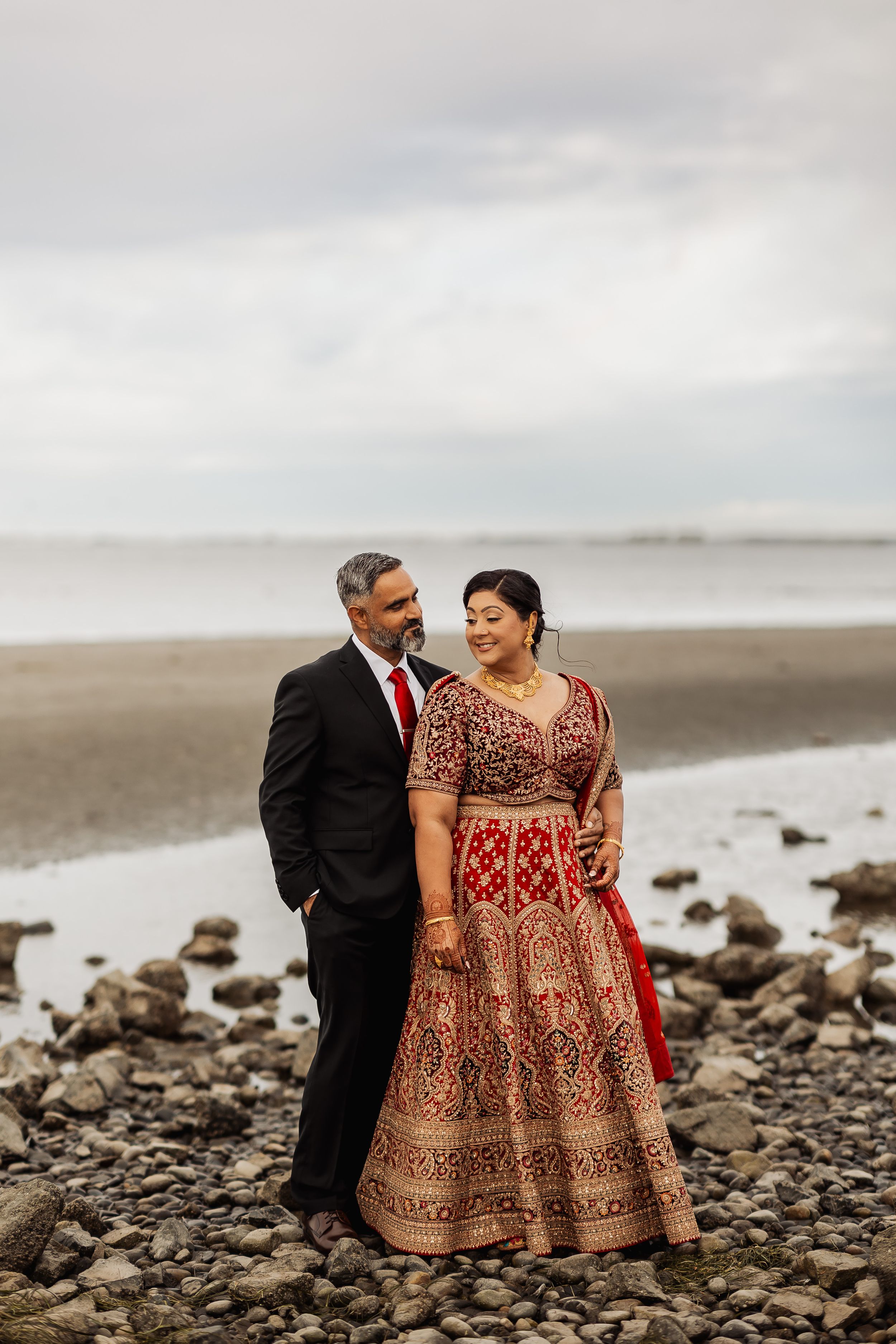 A couple shares romantic moments on a rocky beach shoreline wearing traditional Indian wedding attire.