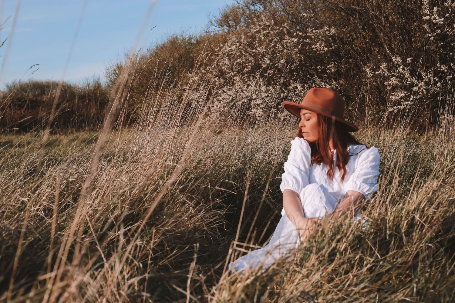 Person in floral dress sitting in tall grass during summer evening playing with flowers.
