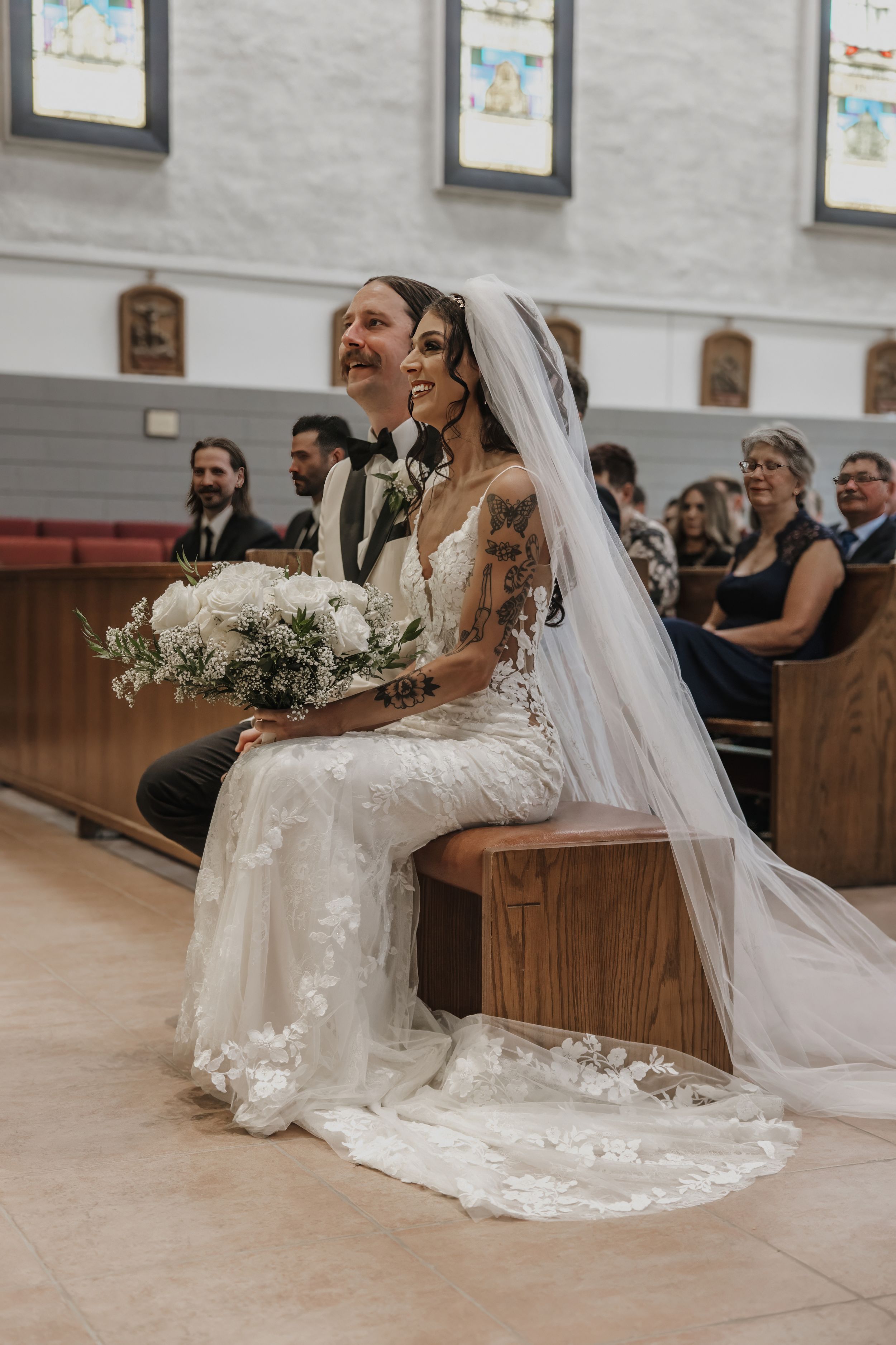 Bride and groom sit together at church altar, bride holding white floral bouquet in lace wedding gown with veil.