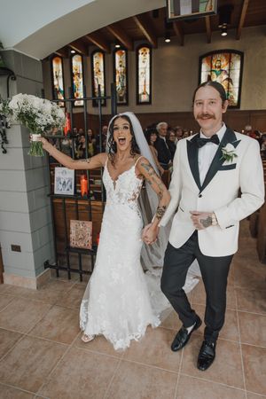Excited bride and groom exit church ceremony, bride raising bouquet in lace gown, groom in white tuxedo jacket.