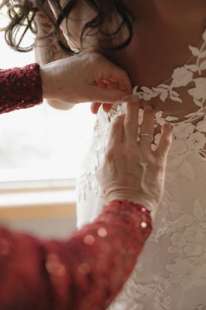 Mother fastening delicate lace buttons on bride's floral wedding gown near a bright window.