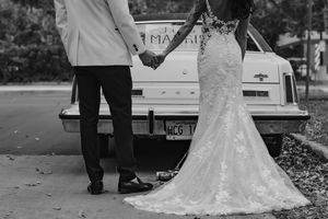 Black and white photo of bride and groom holding hands beside a vintage white getaway car.