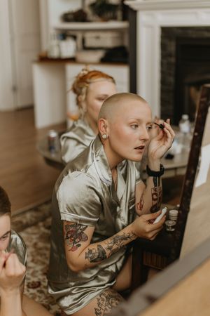 Tattooed bridesmaid with shaved head applying makeup in mirror wearing sage green satin romper.