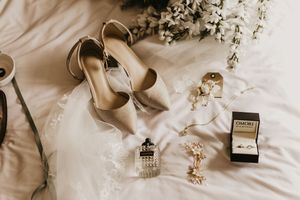Elegant wedding flat lay featuring nude heels, lace bridal gown, perfume, floral accents, and a Pandora ring box on white bedding.