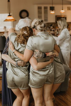 Bridesmaids in matching sage green satin rompers hugging together during wedding morning preparations indoors.