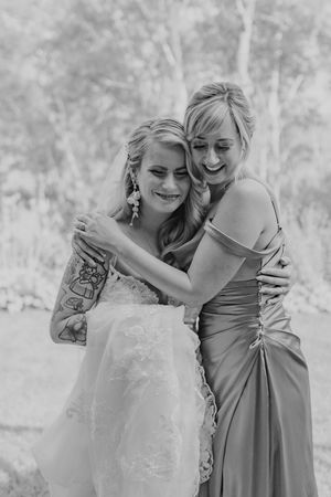Black and white photo of smiling bride in lace gown embracing bridesmaid in satin dress outdoors.