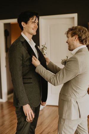 Groom in dark floral suit smiling as groomsman pins boutonniere on lapel during wedding preparations.