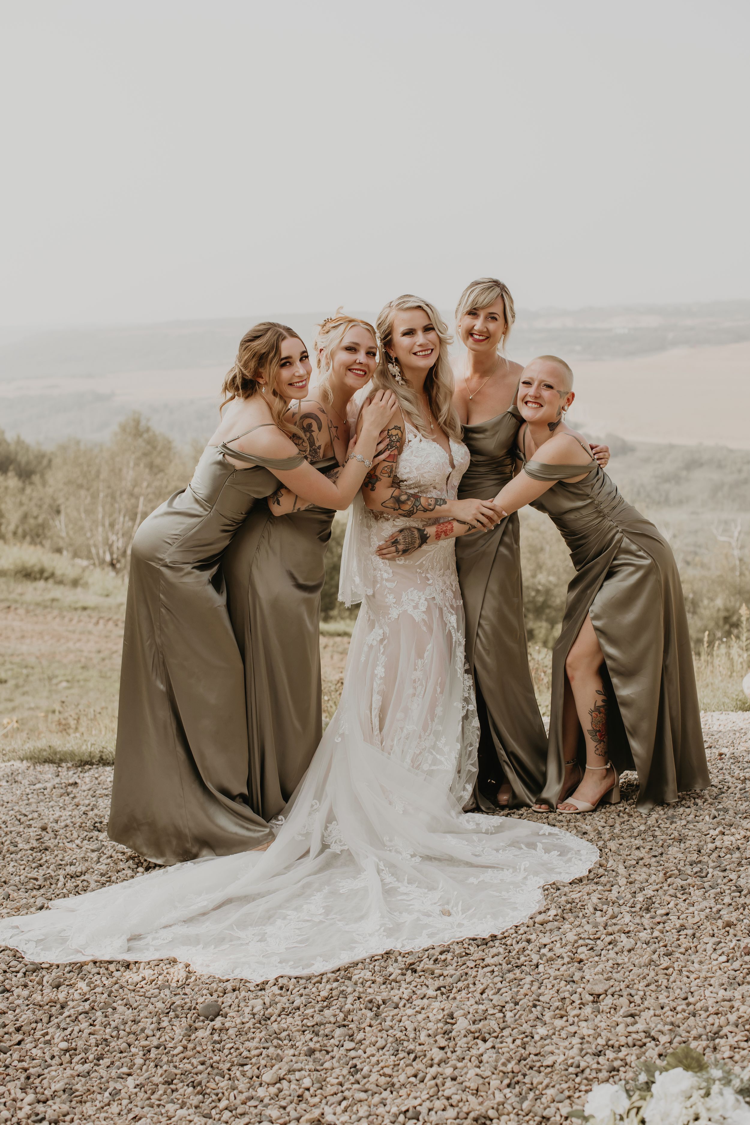 Bride in lace gown surrounded by four bridesmaids in olive green satin dresses with scenic landscape backdrop.