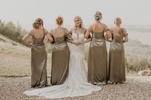 Bride in lace wedding gown posing with four bridesmaids in olive satin dresses viewed from behind outdoors.