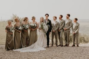 Wedding party posing outdoors with bride in lace gown, groom, bridesmaids in olive dresses, and groomsmen in tan suits.