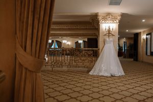 Elegant lace wedding dress hanging in ornate ballroom with grand staircase.