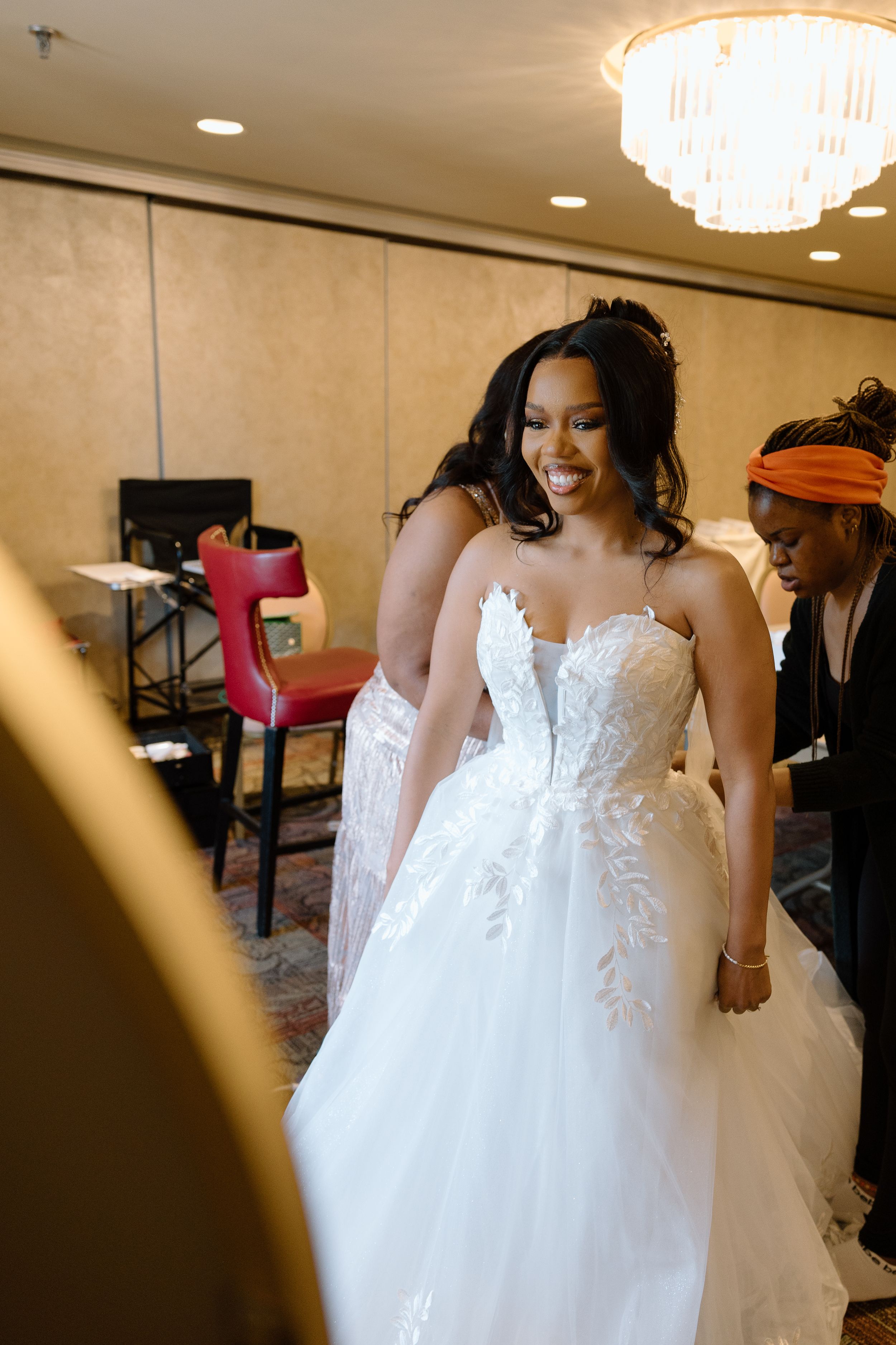 Radiant Black bride in ornate lace ballgown smiles while being assisted in a warmly lit venue.