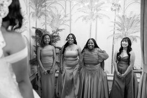 Black and white photo of bridesmaids in gray gowns reacting with joy during bridal first look.