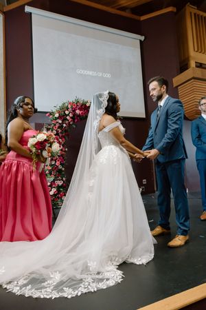 Interracial couple holds hands at church altar during wedding ceremony with floral arch backdrop.