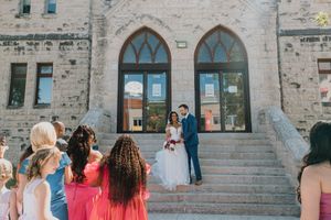 Wedding couple stands on stone church steps surrounded by guests in colorful dresses on a sunny day.