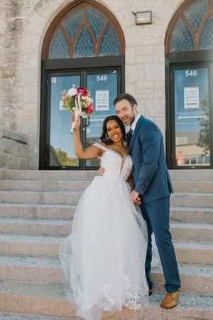 Joyful interracial newlyweds pose on church steps, bride raising colorful bouquet in celebration.