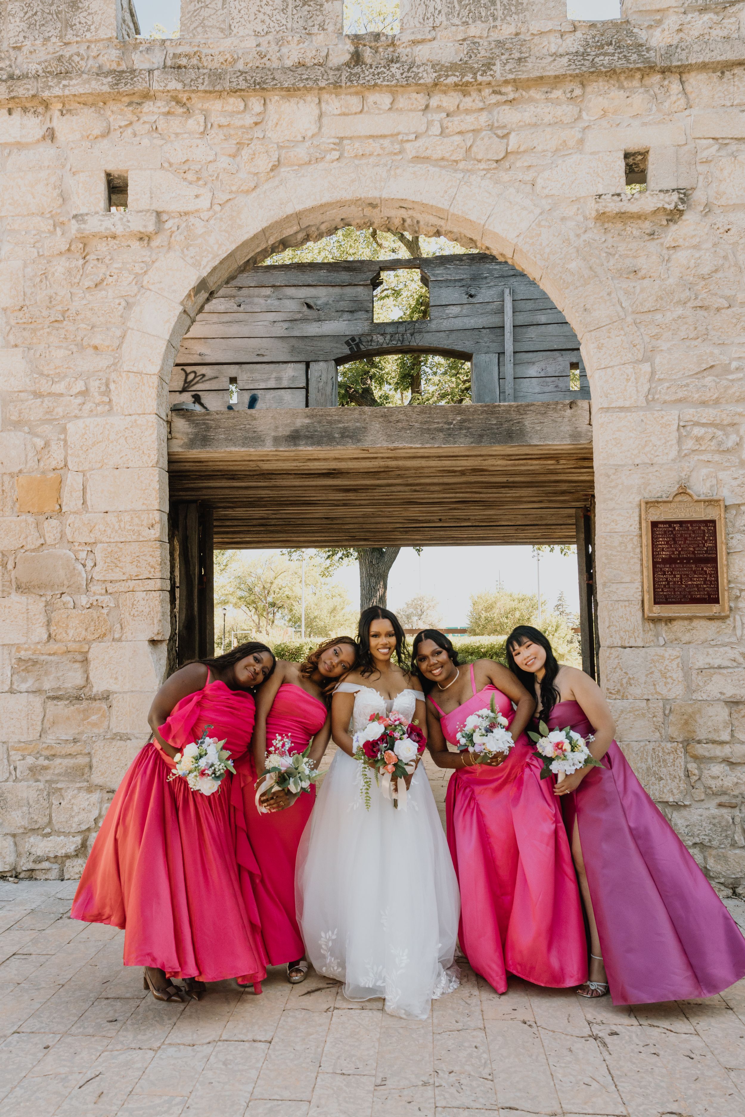 Bride in white gown poses with four bridesmaids in pink ombre dresses holding bouquets under stone archway.