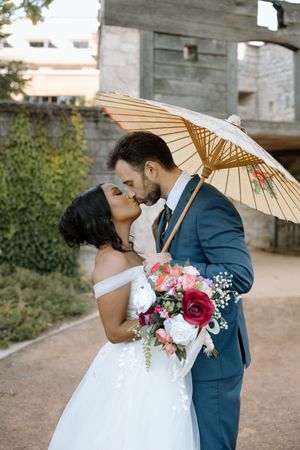 Bride and groom kissing under a decorative parasol umbrella, bride holding a colorful bouquet in a white off-shoulder gown.