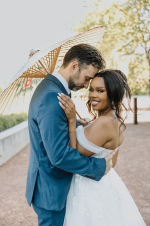 Smiling bride in off-shoulder wedding gown embraces groom in blue suit under a parasol umbrella outdoors.