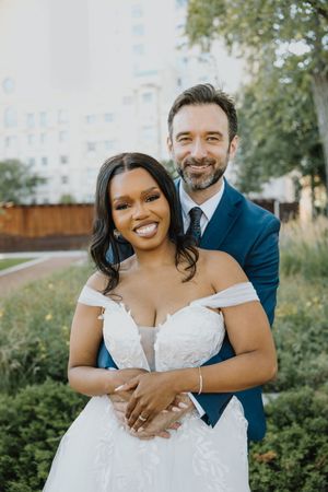Smiling interracial couple posing together outdoors in wedding attire surrounded by greenery.