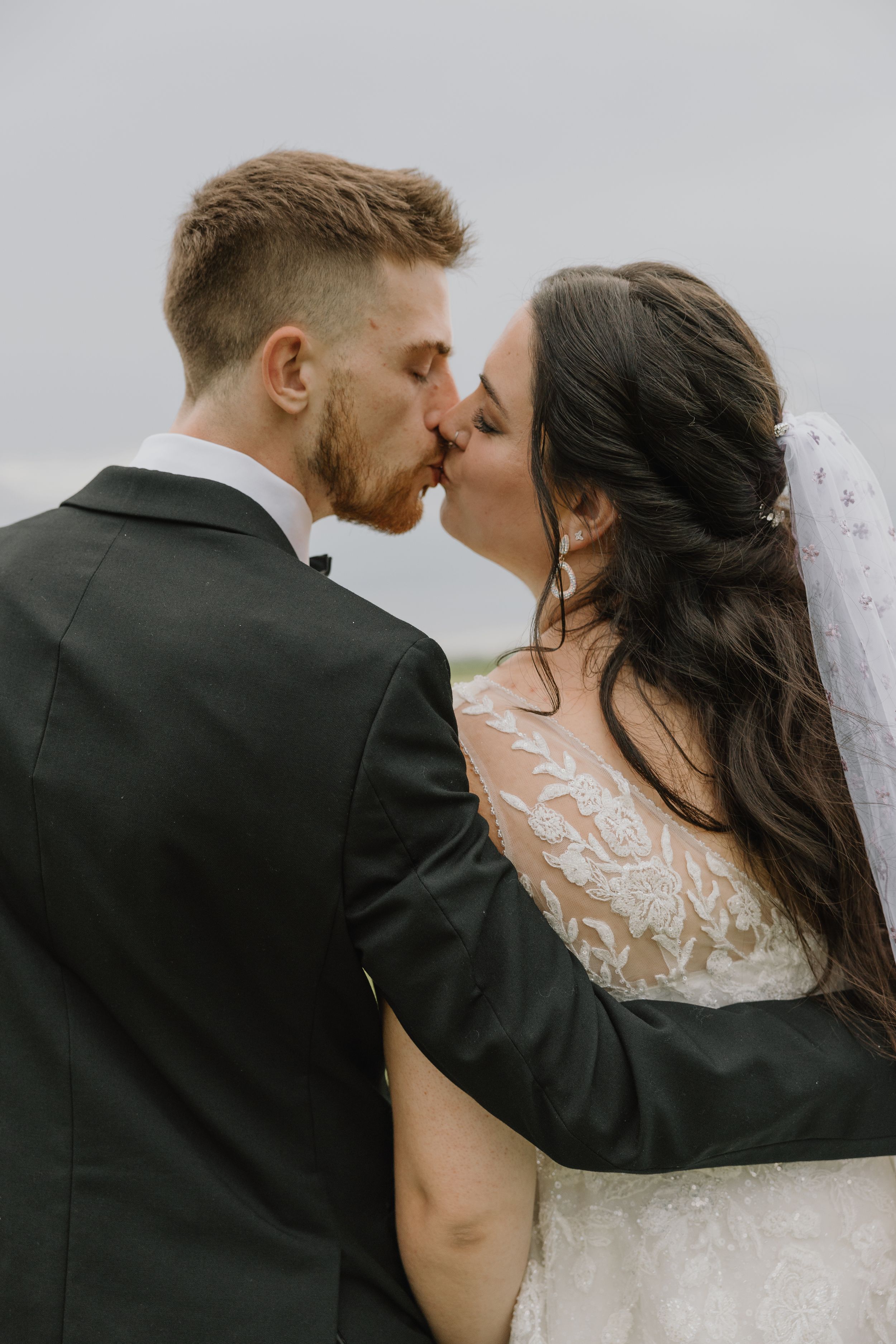 Bride and groom share a romantic kiss outdoors, bride wearing a lace wedding gown with veil.