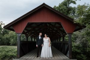 Bride and groom stand together under a red covered bridge surrounded by lush green trees.