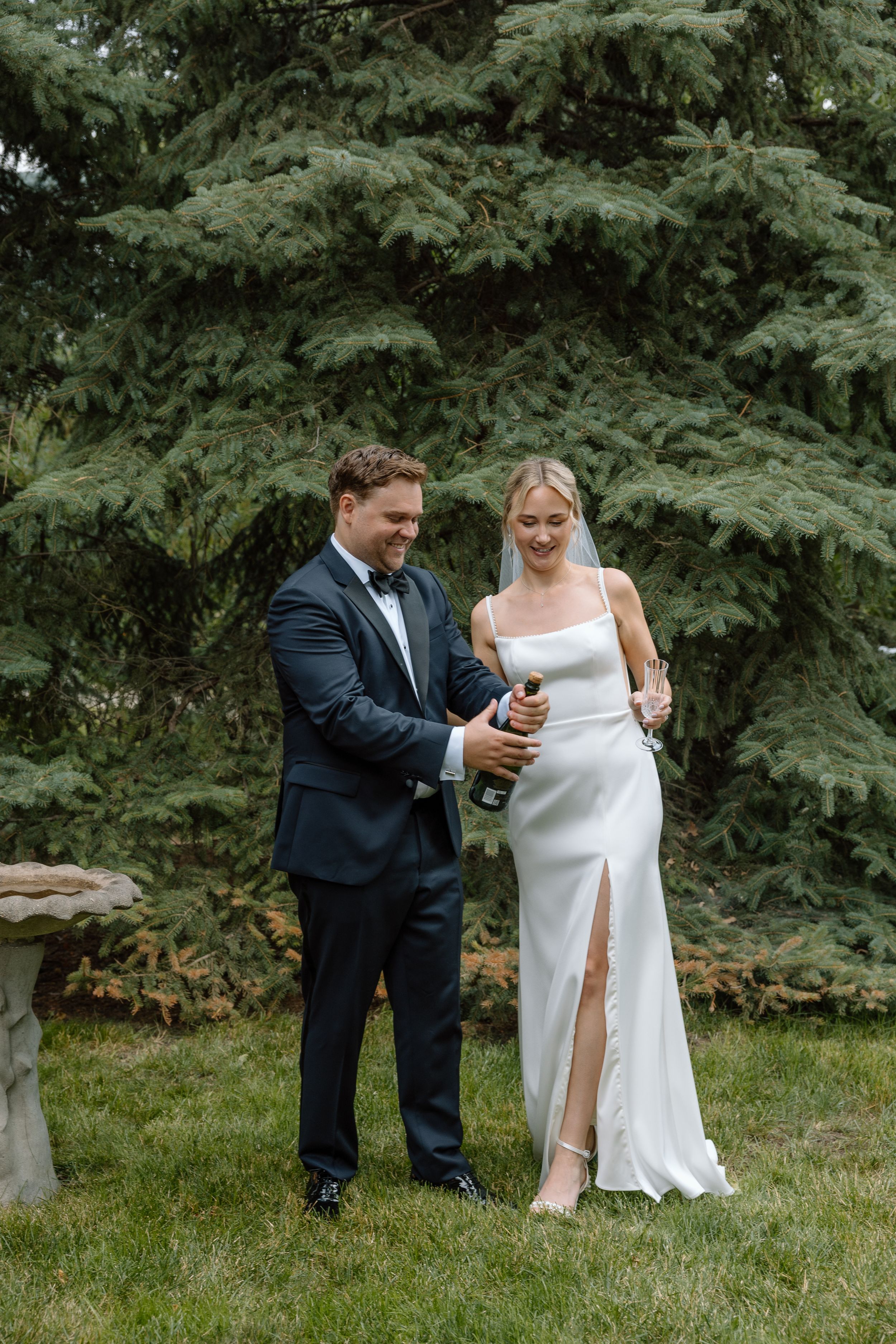 Groom in navy tuxedo opens champagne bottle beside smiling bride in sleek white gown outdoors.