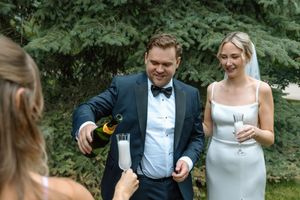 Groom in navy tuxedo pours champagne for a guest while bride in white dress smiles nearby.