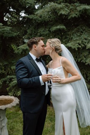 Bride and groom kissing while holding champagne flutes outdoors near evergreen trees on their wedding day.