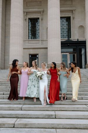 Bride with white bouquet walks down monument steps flanked by six bridesmaids in colorful mismatched gowns.