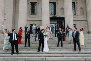 Wedding party celebrates on grand monument steps as bride and groom kiss surrounded by cheering guests.