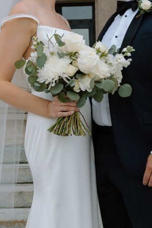 Bride holding elegant white rose and eucalyptus wedding bouquet while standing with groom in black tuxedo.