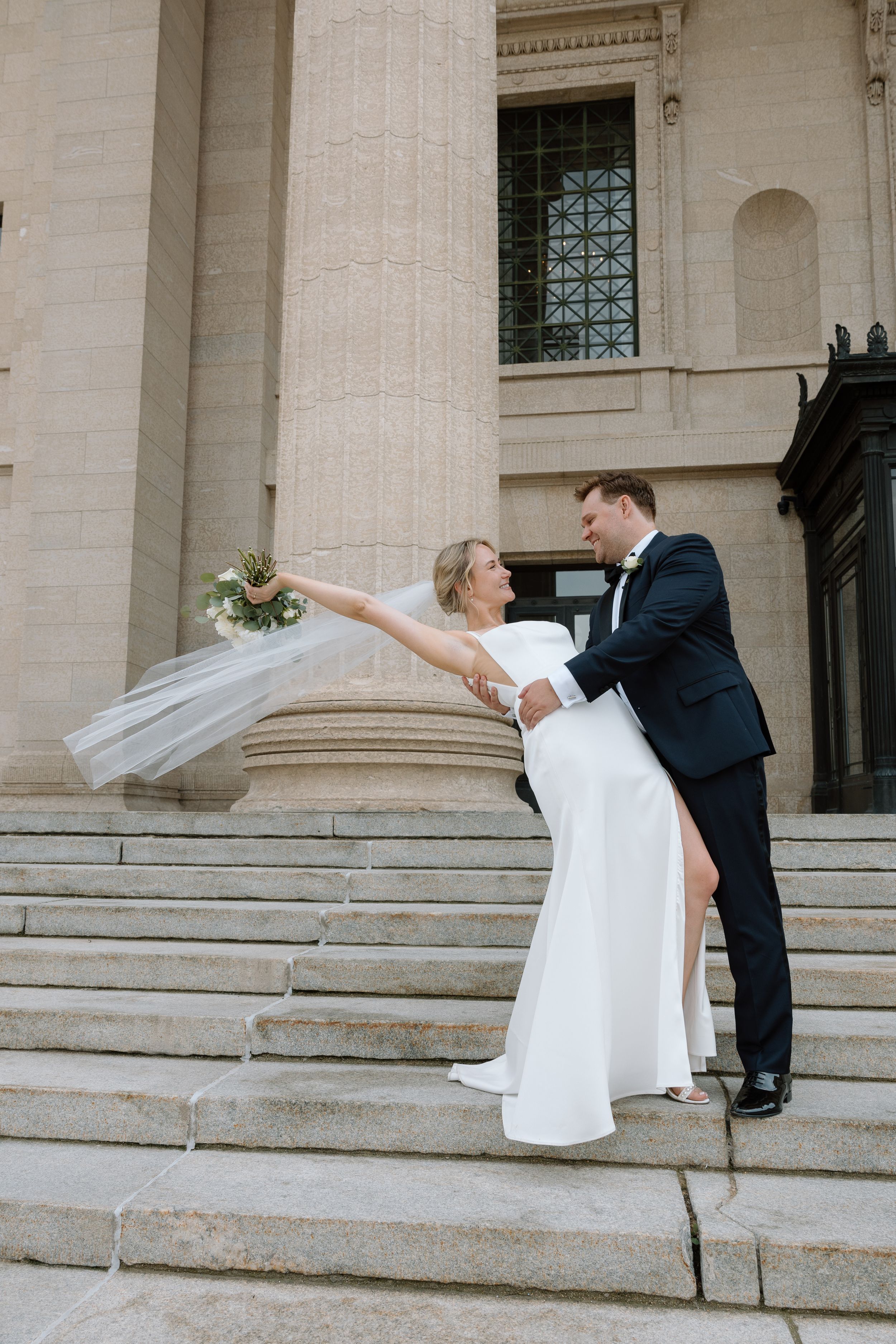 Groom dipping bride on grand stone staircase as her veil flows in the wind during wedding portraits.