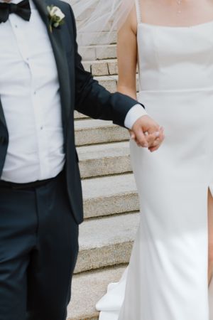 Bride and groom holding hands while walking down stone steps, bride in sleek white gown with veil.