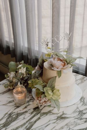 Two-tier white wedding cake with floral decorations and heart topper on marble table.