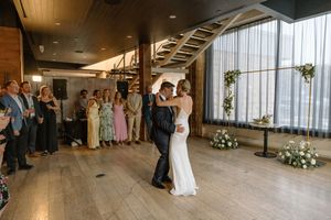 Bride and groom share first dance on wooden floor surrounded by wedding guests in modern venue with large windows.