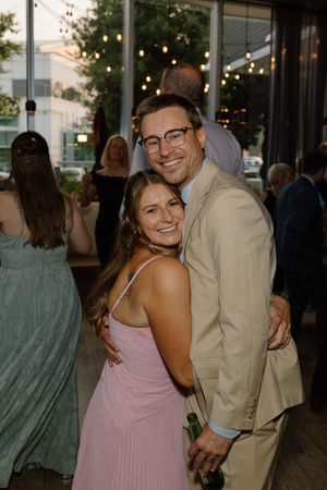 Smiling couple embraces at outdoor wedding reception, man in tan suit and woman in pink dress under string lights.