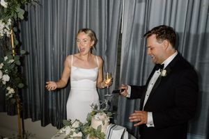 Bride in white gown and groom in tuxedo stand at wedding cake table with champagne glasses and floral arrangements.
