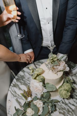 Bride and groom cutting white wedding cake decorated with flowers on marble table.