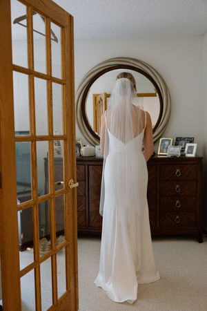 Bride in a simple white gown and veil viewed from behind standing before a round mirror.