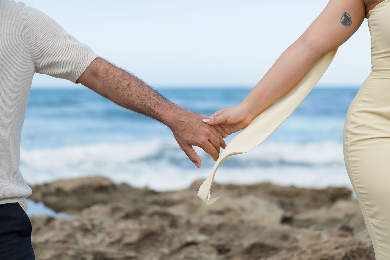 A bride and groom hold hands on a beach, with the ocean in the background.