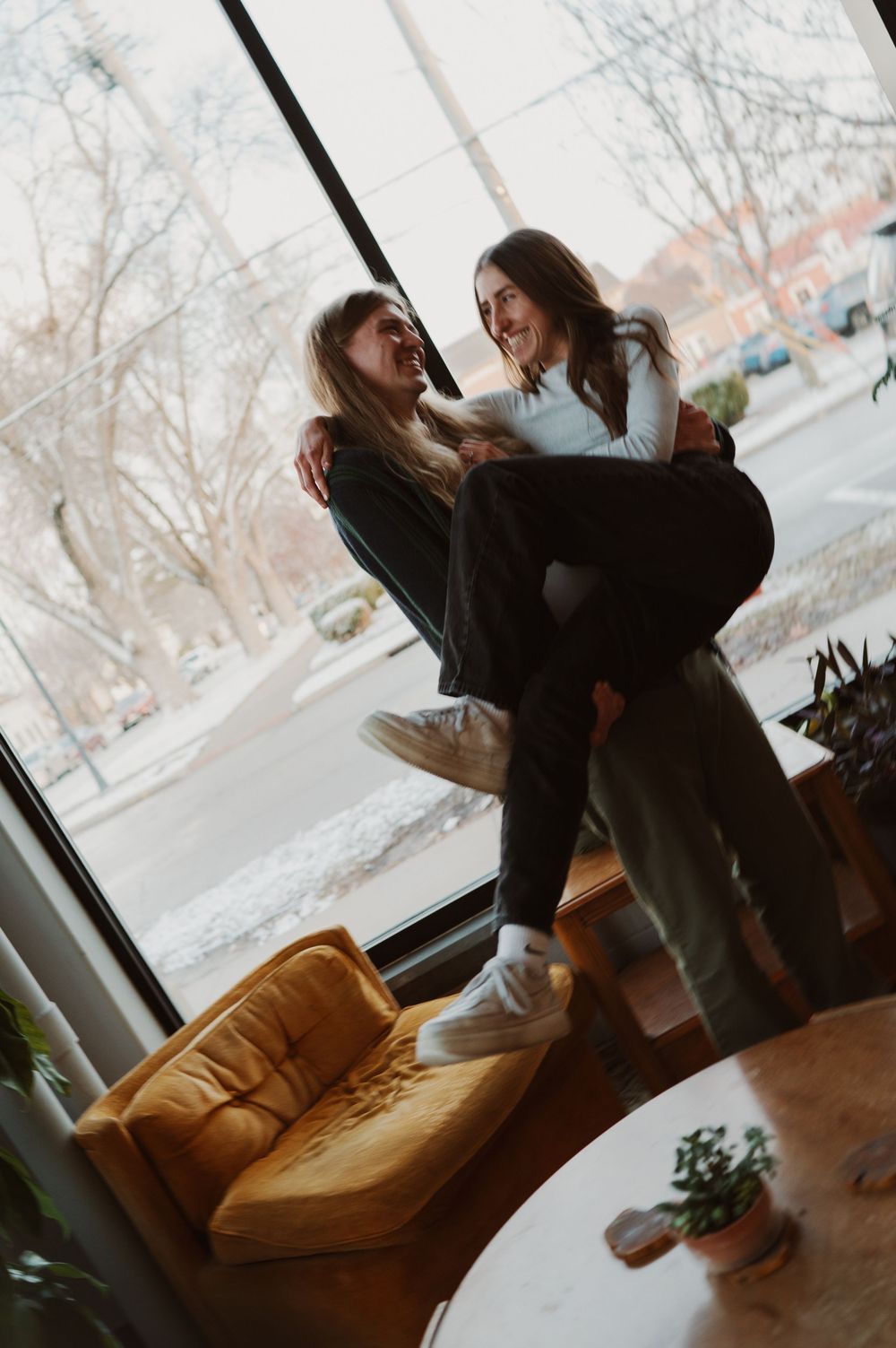 Two friends laugh and play together near a large window with snowy winter scene outside, next to tan leather couch.