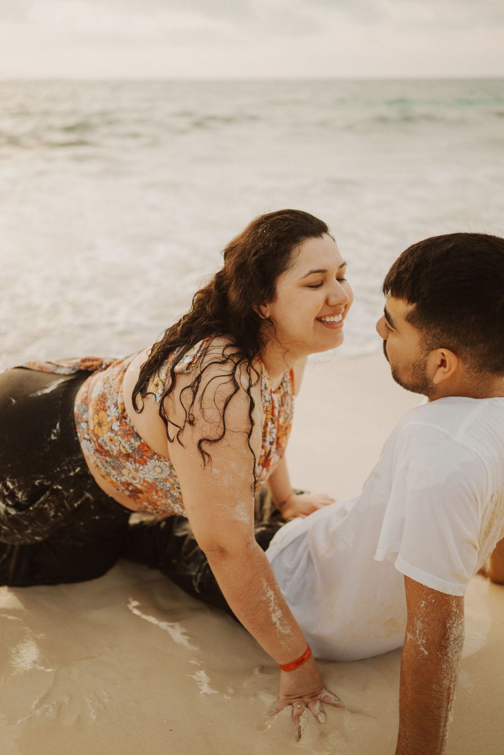 A playful couple laughs together while sitting on a beach at sunset.
