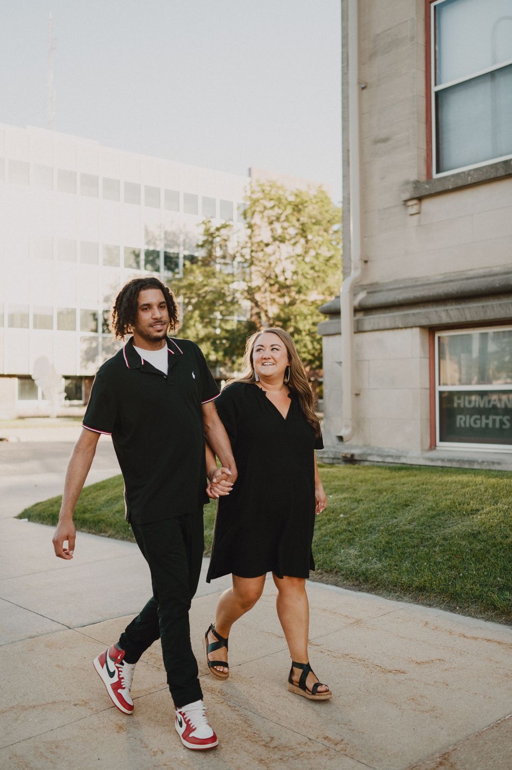 A couple wearing matching black outfits holds hands while walking together on a sidewalk on a sunny day.