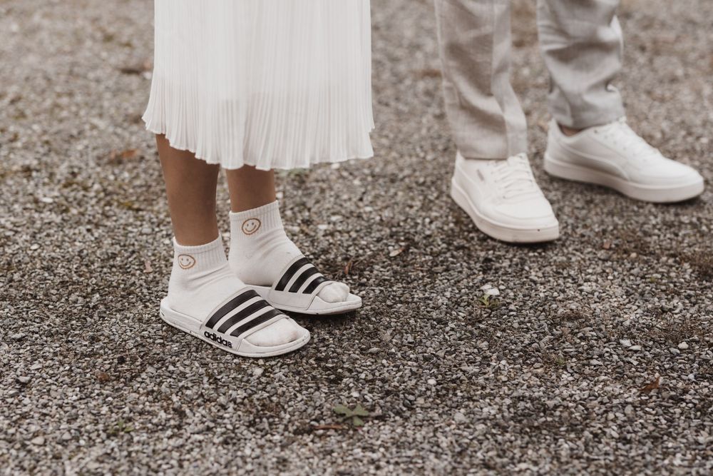 Nahaufnahme von Hochzeitspaar in Adidas-Turnschuhen und Adiletten auf Schotter, im Kontrast zu heller Kleidung.