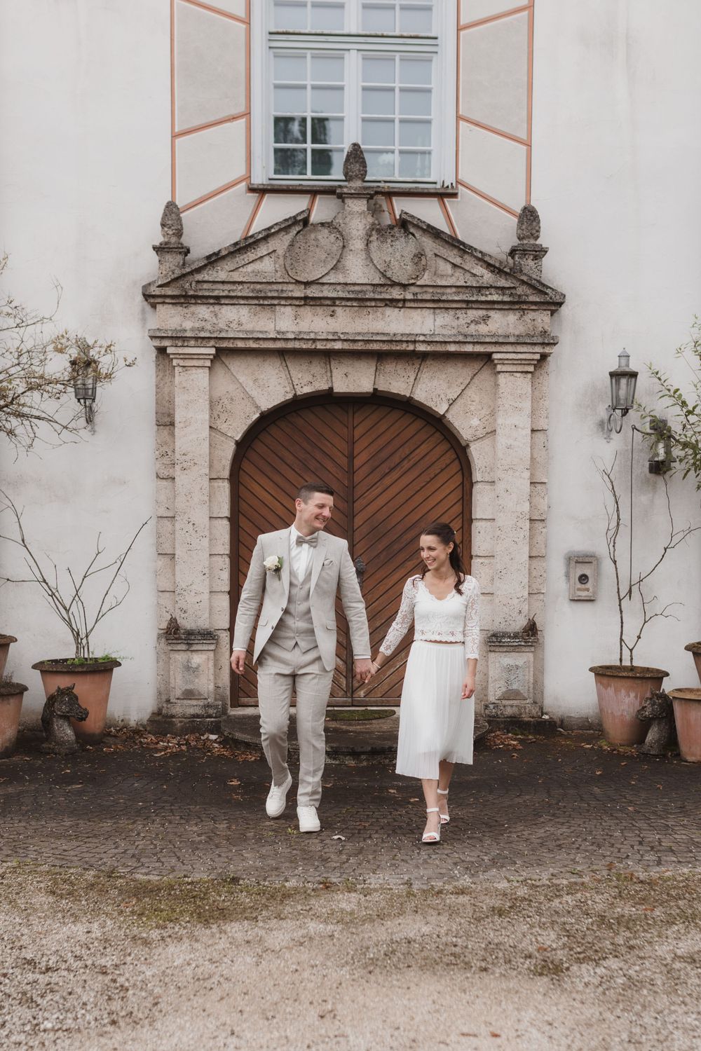 Zwei Menschen in Hochzeitskleidung gehen Hand in Hand durch einen reich verzierten Steintorbogeneingang eines historischen Gebäudes.