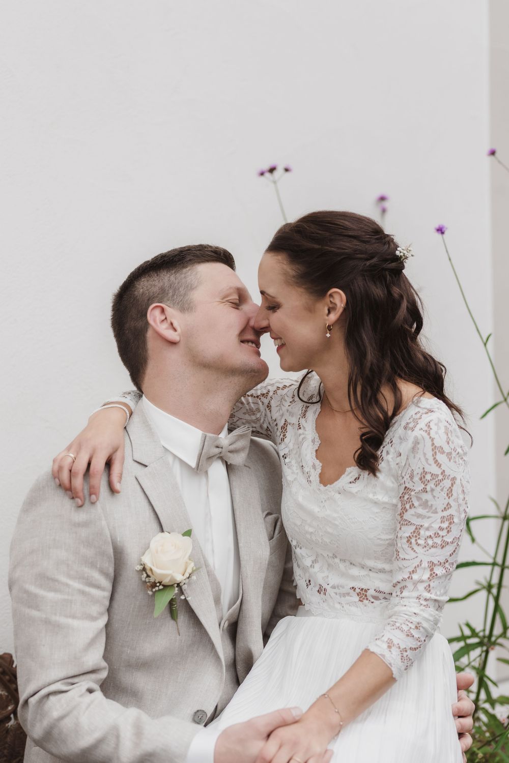 Ein zärtlicher romantischer Moment zwischen Frischvermählten in weißer Spitze und beigem Gewand vor einem hellen Hintergrund.